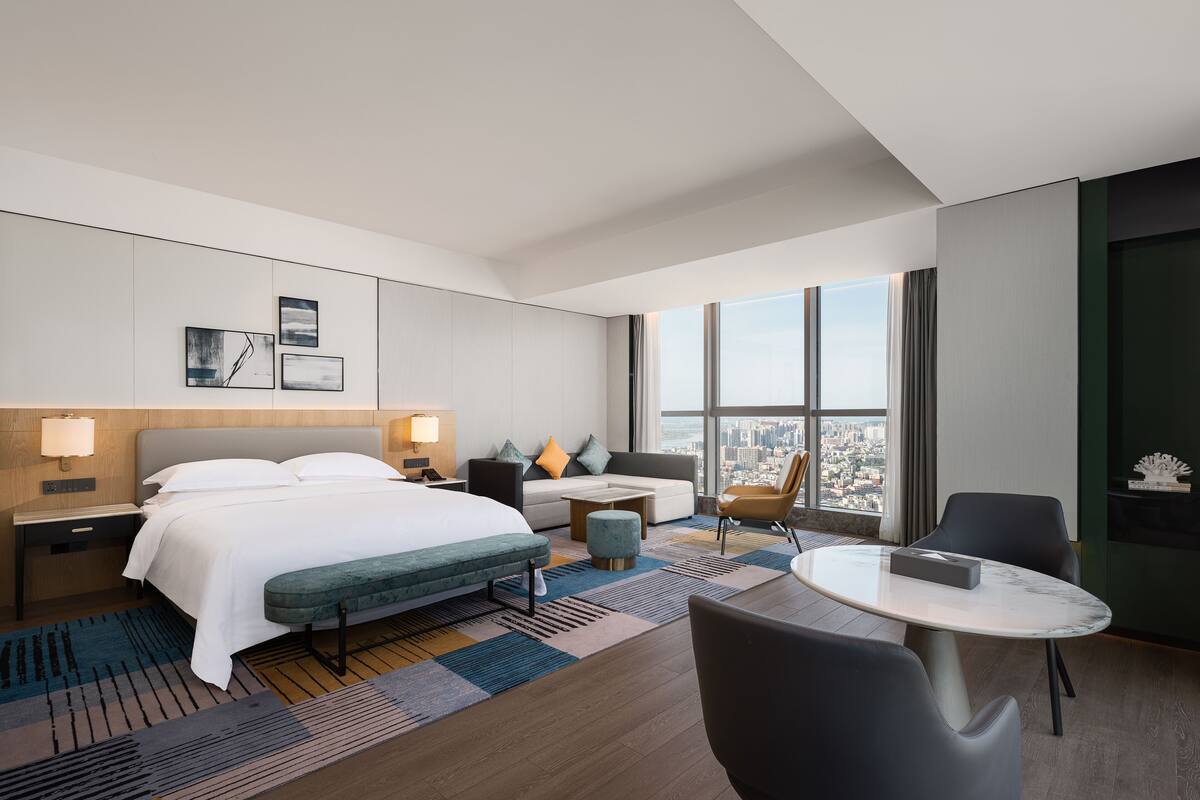A modern hotel room with a large bed, a sofa, a small table, and a view of the city.