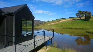A modern cabin with a large glass window, a deck, and a view of a lake and hills. - Beautifully appointed Tiny House. (Goughs Bay)