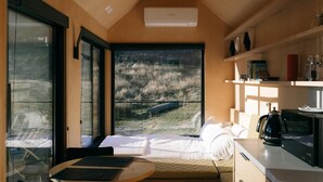 A bedroom with a large window, a bed, a bedside table, a microwave, and a shelf with books and decorative items. - Beautifully appointed Tiny House. (Goughs Bay)