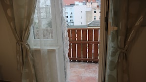 A view from a window with curtains tied back, revealing a balcony with a wooden fence and buildings in the background. - Penthaus Am Köpcke (Hannover)