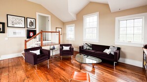 A spacious living room with hardwood flooring, a dark-colored sofa, two armchairs, a coffee table, and a staircase leading to an upper level. - Chef’s Kitchen, Fast Wi-Fi (Hillsdale)