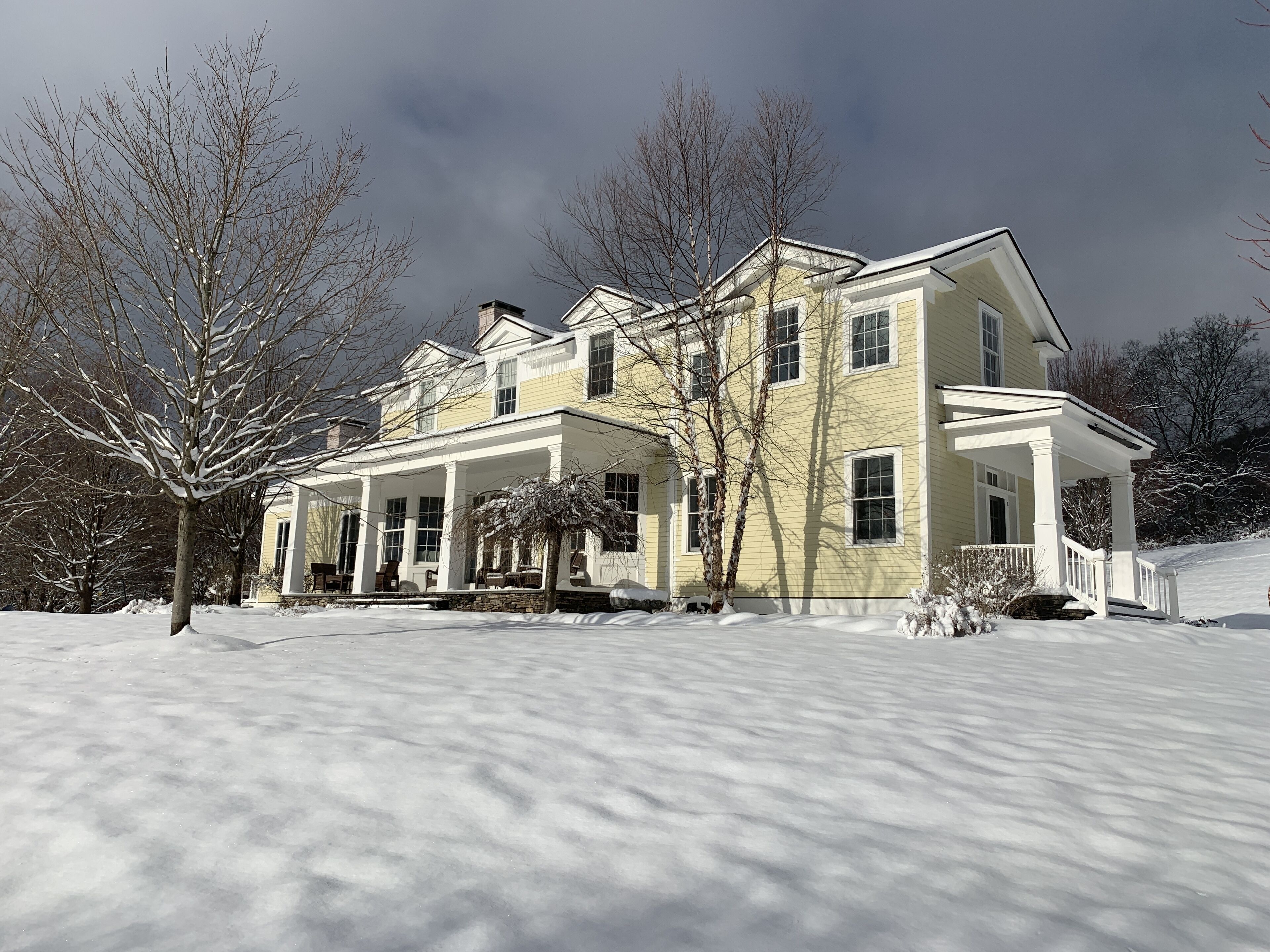Una casa grande de dos pisos con un porche cubierto y un jardín lleno de nieve.
