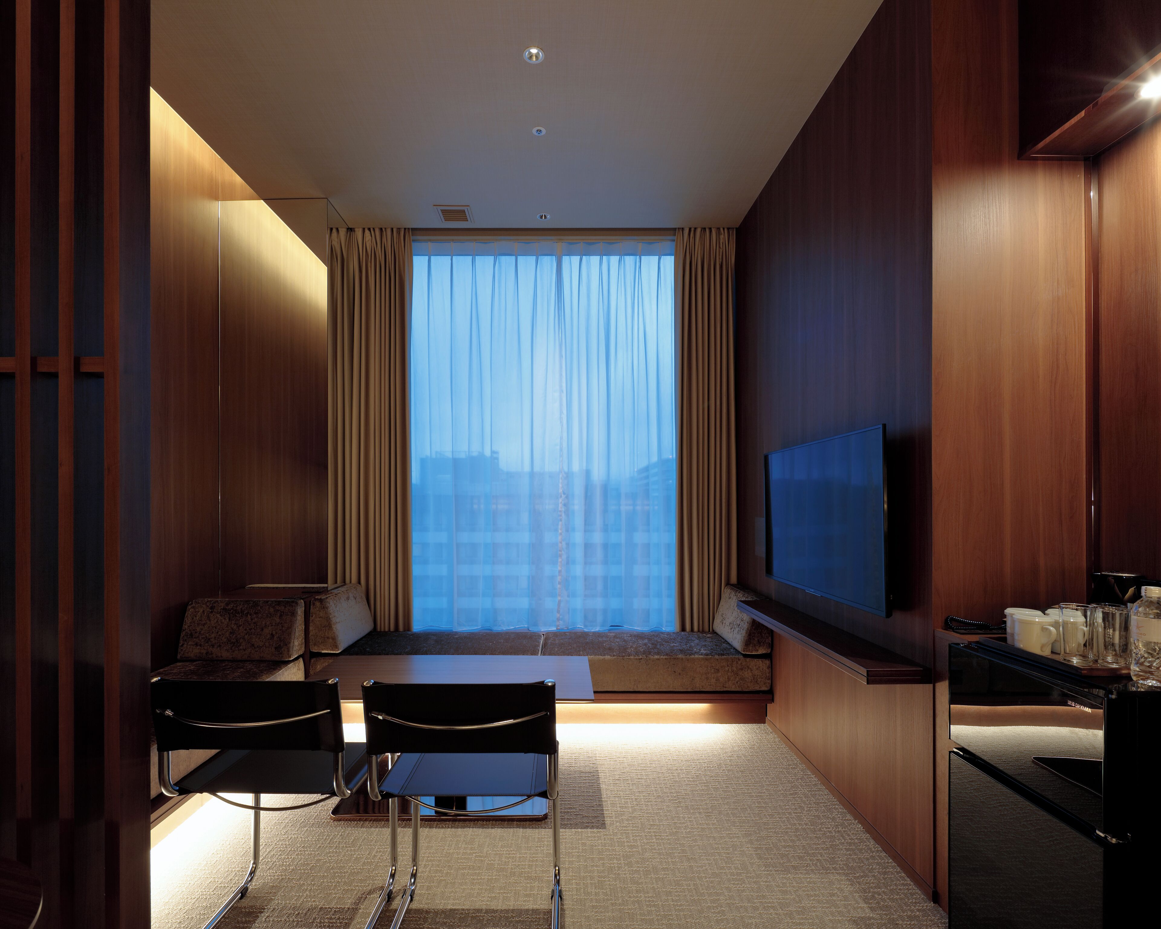 Een moderne hotelkamer met een groot raam, een flatscreen televisie, een bank, een eettafel en een minibar.