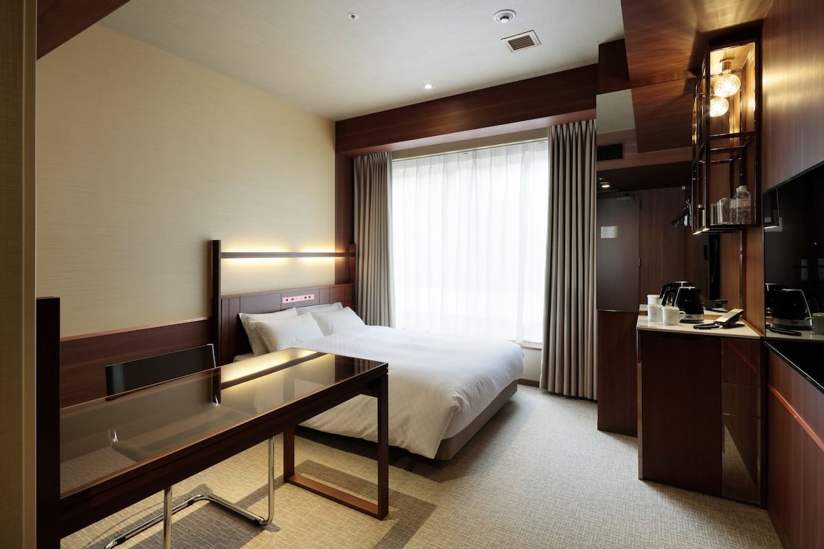 A hotel room with a bed, bedside table, and a desk with a chair. There is a window with curtains, a television, and a minibar.
