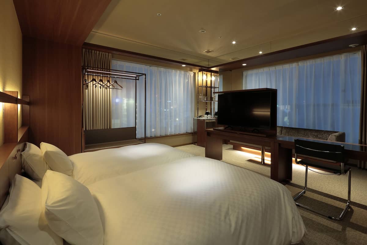 A hotel room with a large bed, a TV, a desk, and a chair. There is a window with curtains and a view of the city.