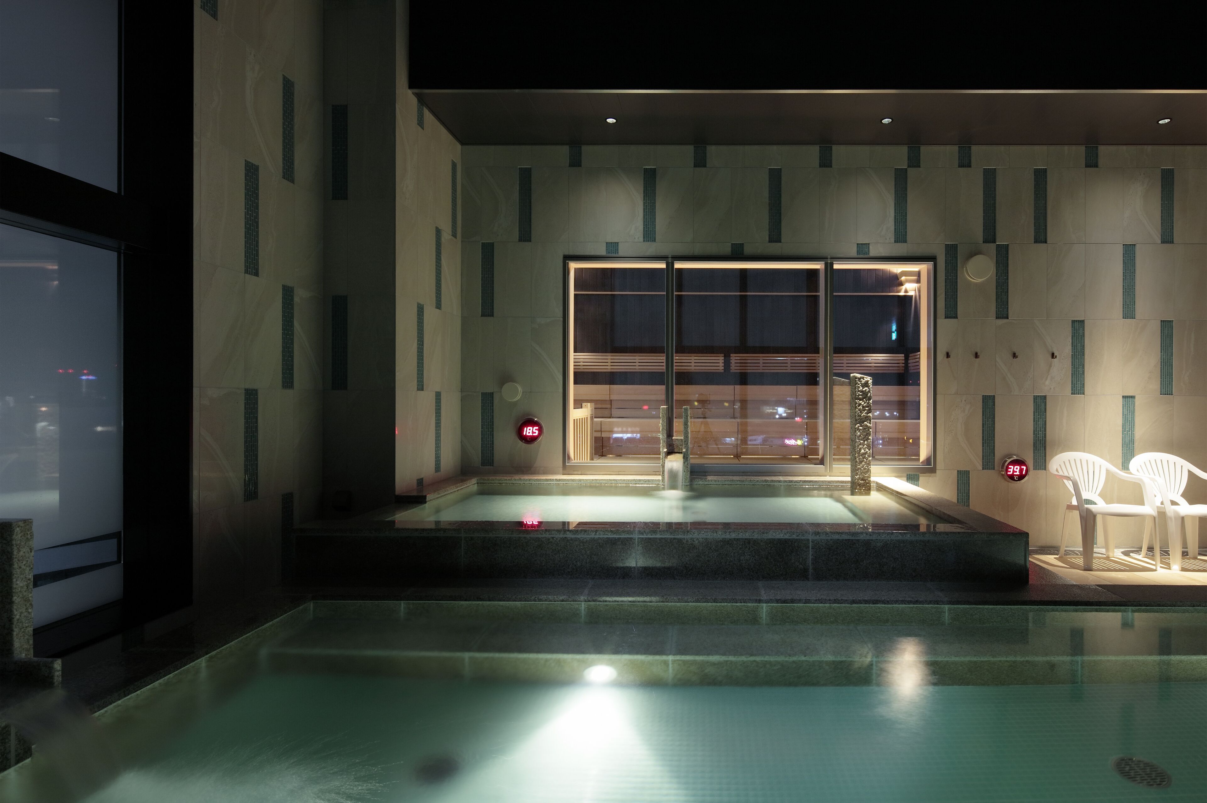 A modern indoor pool area with a glass enclosure, steps leading into the pool, and two white chairs by the poolside.