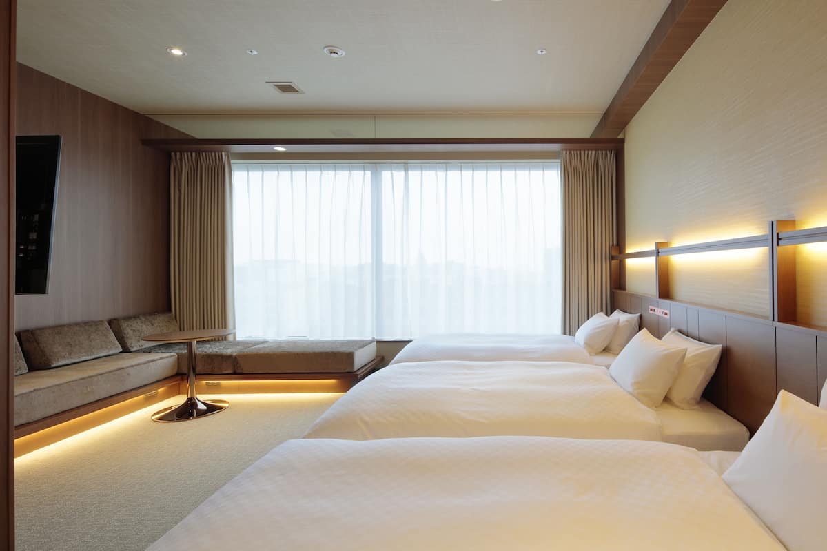 A hotel room with two beds, a sofa, a small table, and a large window with curtains.