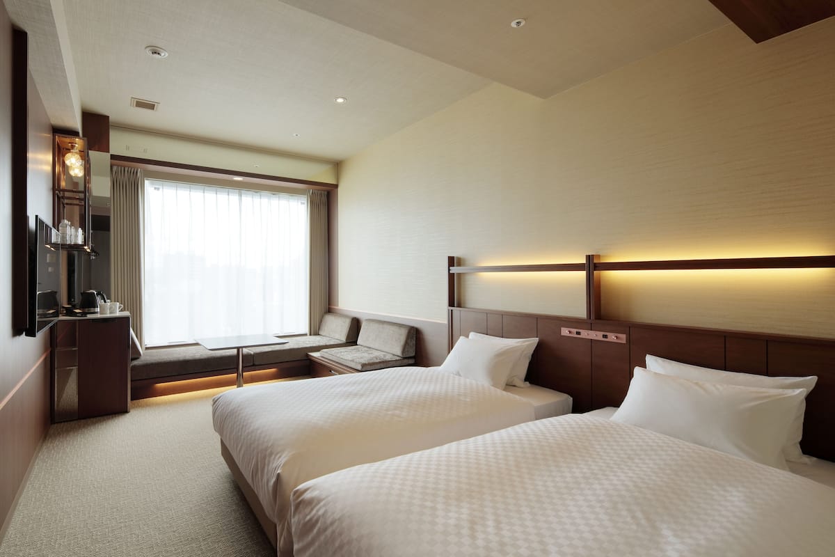 A hotel room with two beds, a desk, a chair, a TV, and a window with curtains.