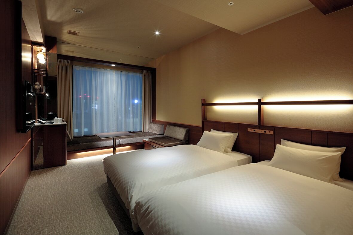 Habitación de hotel con dos camas, un escritorio, una silla, un ventanal con cortinas y una mesita de noche.