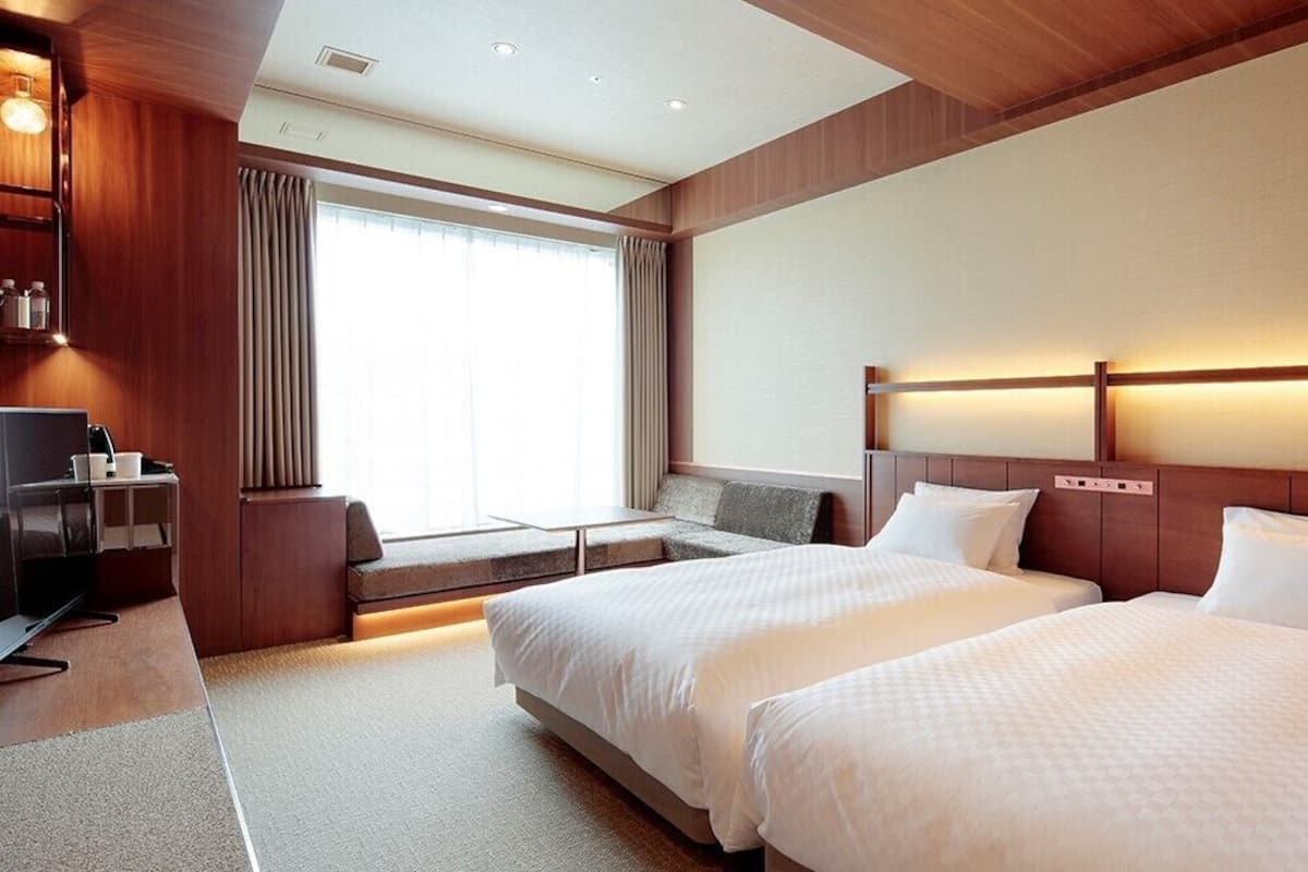 A hotel room with two beds, a desk, a TV, and a large window with curtains.