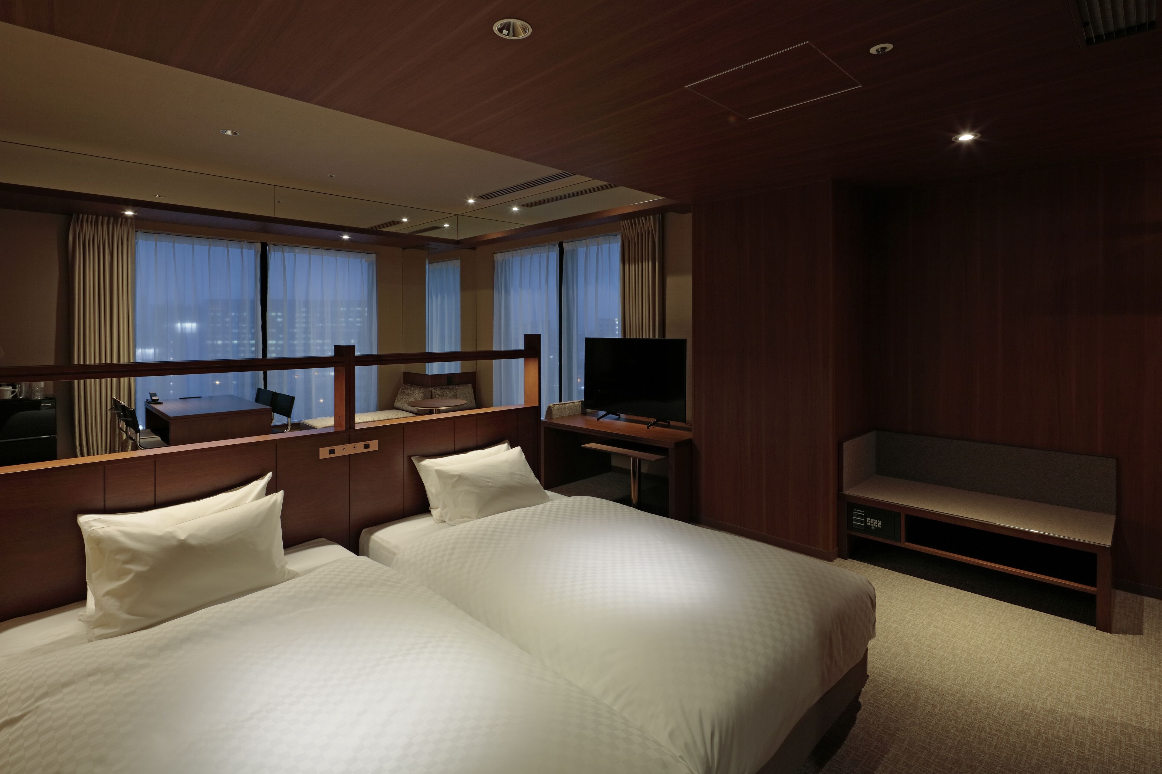 Quarto de hotel com uma cama grande, uma escrivaninha, uma televisão e vista para a cidade através das janelas.