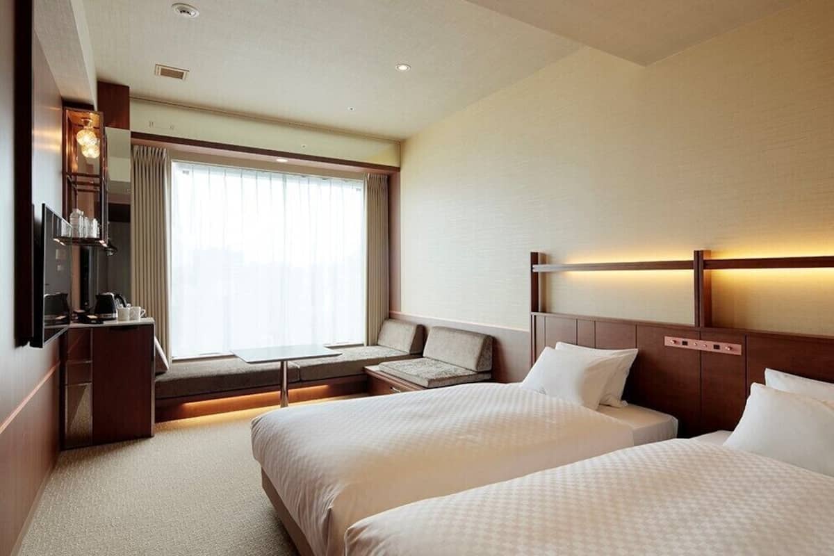 A hotel room with two beds, a desk, a TV, and a window with curtains.