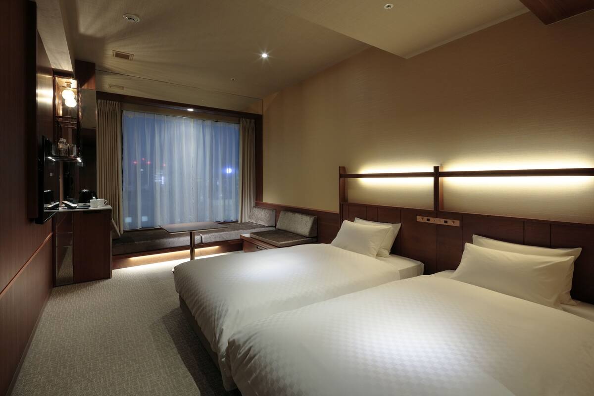 A hotel room with two beds, a desk, a chair, and a window with curtains.