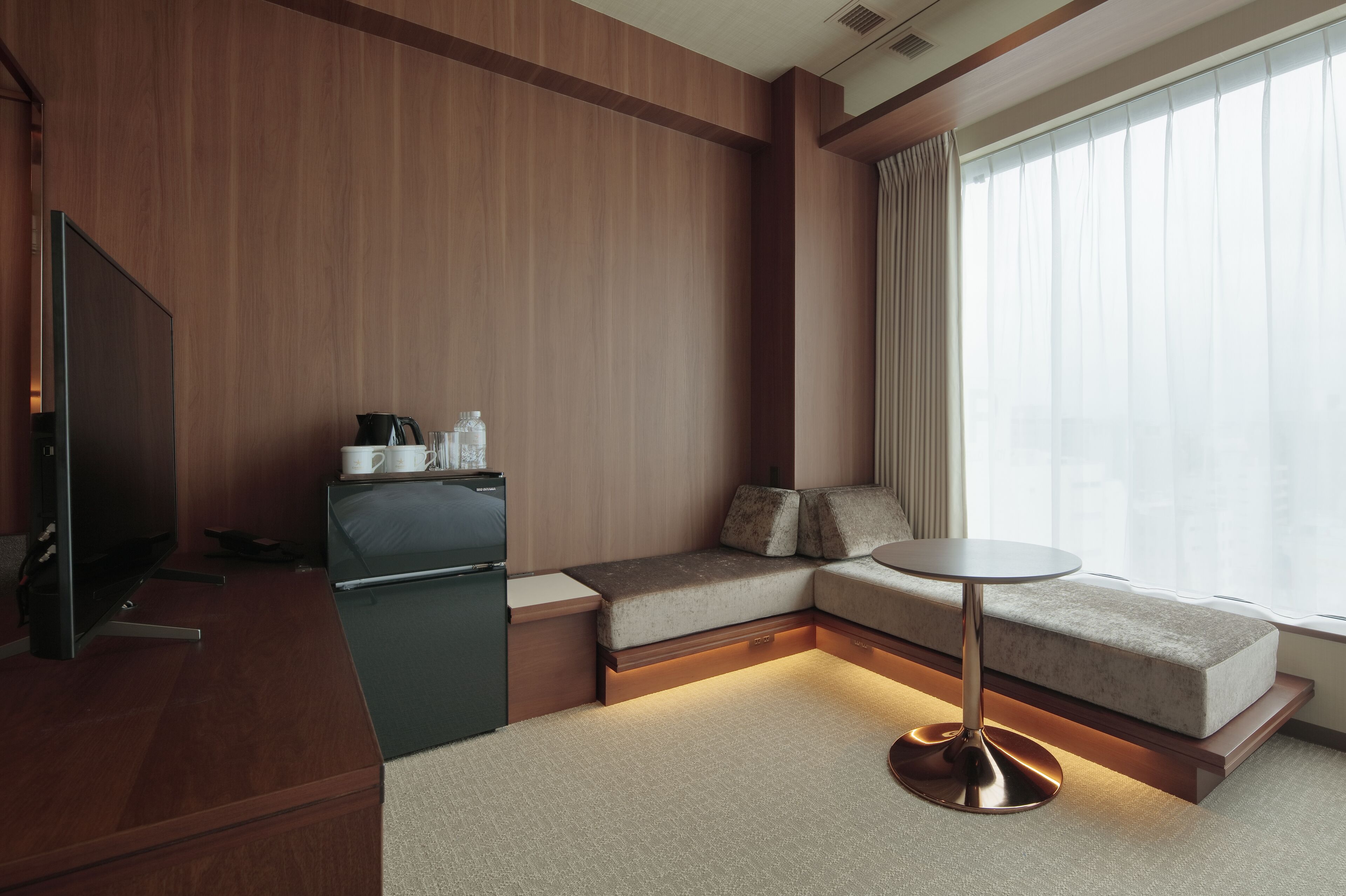 A modern hotel room with a wooden panel wall, a sofa, a small table, a television, and a window with curtains.