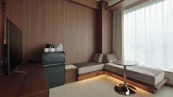 A modern hotel room with a wooden panel wall, a sofa, a small table, a television, and a window with curtains.
