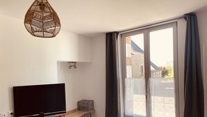 A modern room with a flat-screen TV on a wooden TV stand, a desk with a chair, a large window with curtains, and a hanging light fixture.