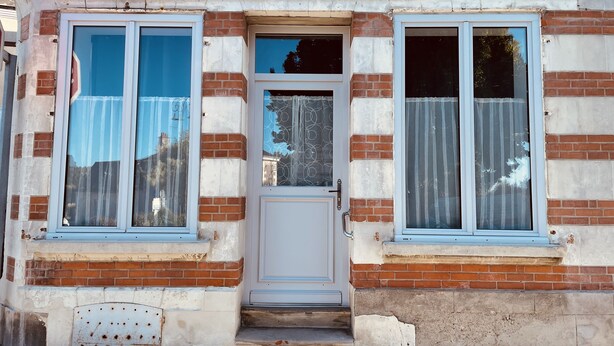 A two-story building with a brick facade, a central door, and two windows on the ground floor.