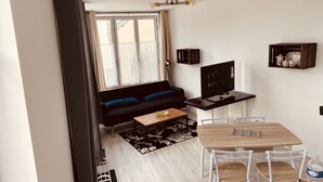 A modern living room with a black sofa, a wooden coffee table, a TV on a stand, and a dining area with a wooden table and chairs.