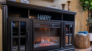 Une cheminée noire avec une porte en verre, une inscription au-dessus qui dit « JOYEUX », et un panier en osier avec un coussin posé sur le sol à côté.