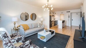 Een moderne woonkamer met een grijze bank, een fauteuil met patroon, een salontafel, en een eethoek met een keuken op de achtergrond.