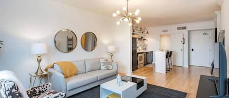 Una sala moderna con un sofá gris, un sillón estampado, una mesita de centro y un comedor con cocina al fondo.