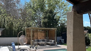 A covered outdoor area with a pool, lounge chairs, a wooden deck, and a pergola with a woven roof.