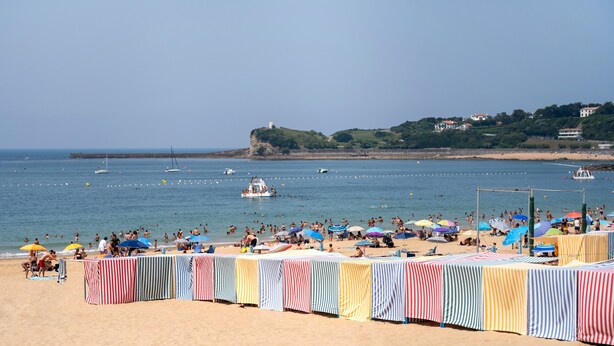Une plage avec des auvents rayés, un rivage sablonneux et des bateaux sur l’eau.