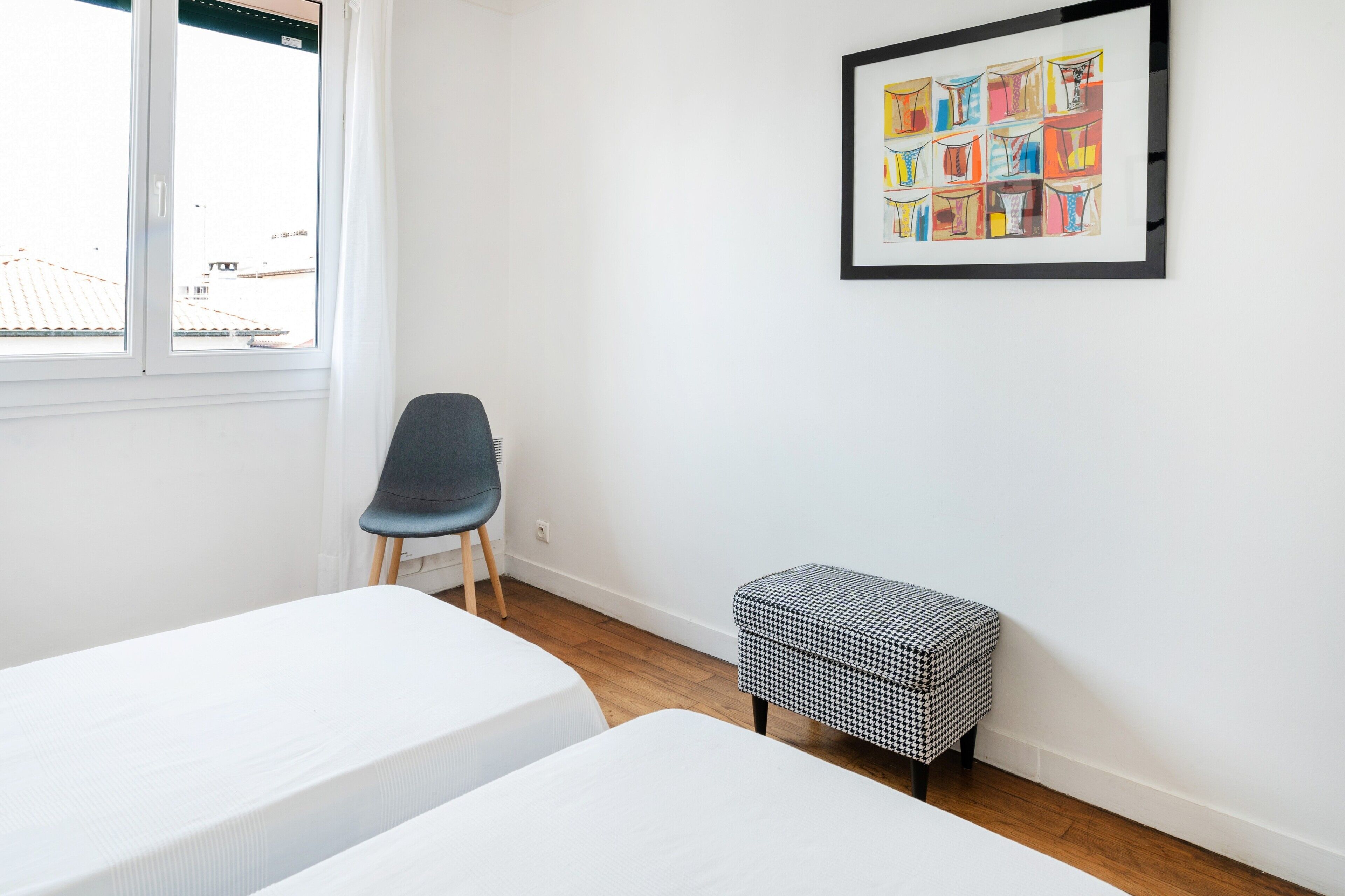 A room with two beds, a chair, a small bench, a window, and a framed artwork on the wall.