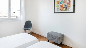 A room with two beds, a chair, a small bench, a window, and a framed artwork on the wall.