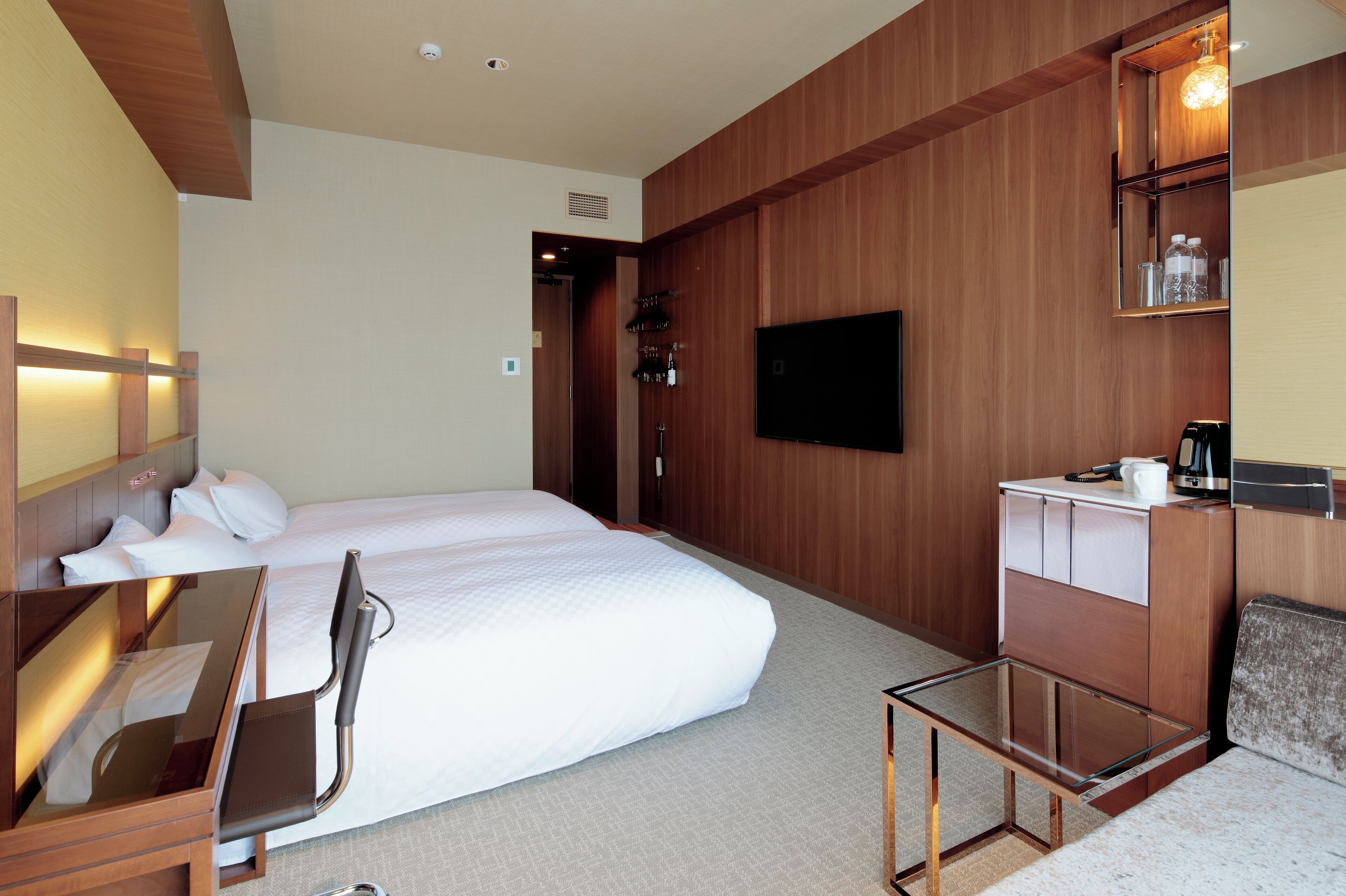 A hotel room with a bed, a desk with a chair, a TV, a minibar, and a small table.