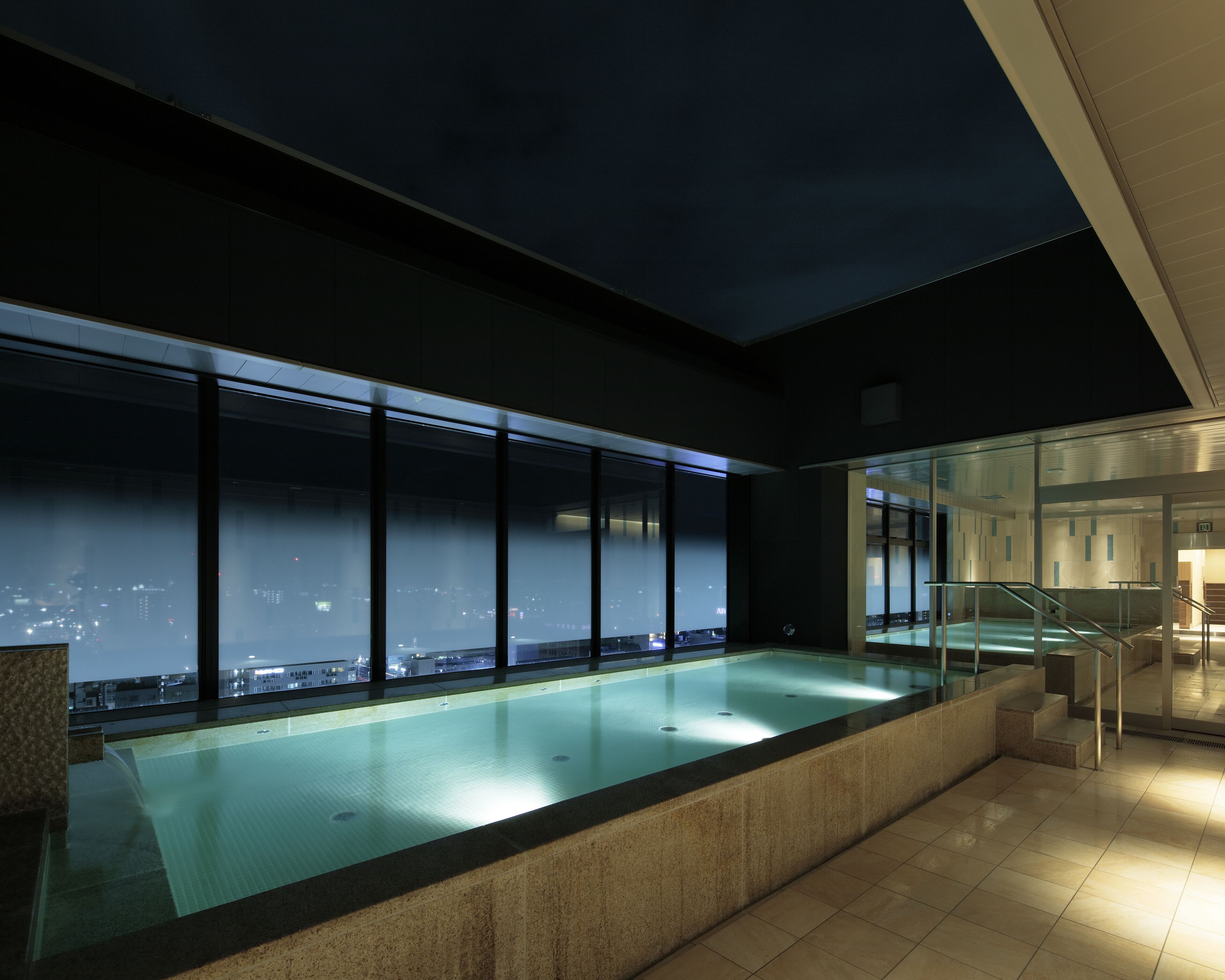 A modern indoor pool with a city view, large windows, and a staircase leading to an upper level.