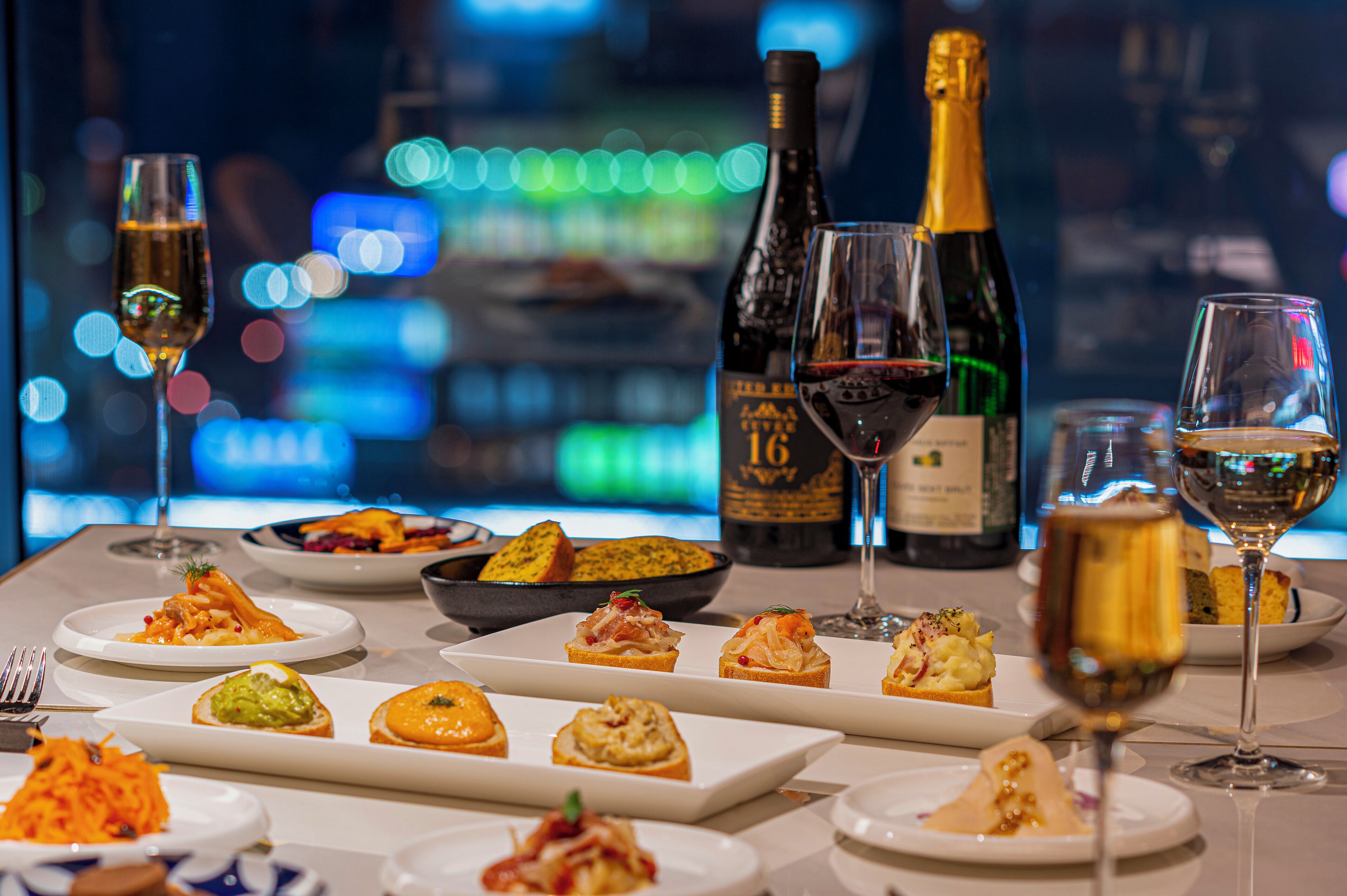 A table set with various dishes and glasses of wine, with a cityscape view in the background.