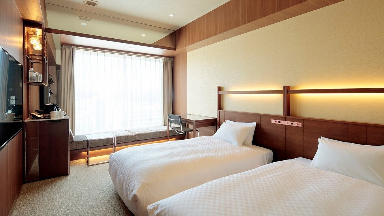 A hotel room with two beds, a desk, a chair, a TV, and a large window with curtains.