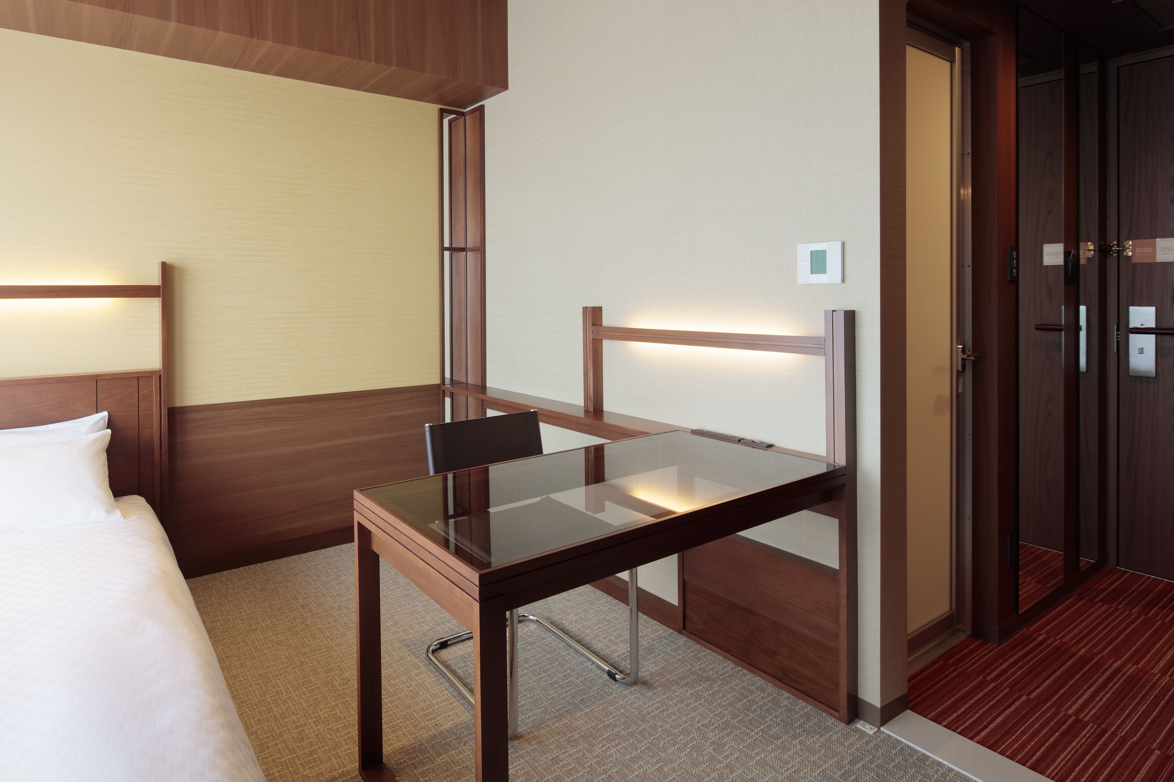 A hotel room with a bed, a glass-top table, a chair, a wooden nightstand, and a door.