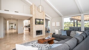 Een ruime woonkamer met een grote grijze modulaire bank, een houten salontafel en een stenen open haard. Achtergrond is een keuken en er is een balkon met uitzicht op groen.