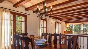 Un comedor con mesas y sillas de madera, una araña y ventanas grandes con cortinas.