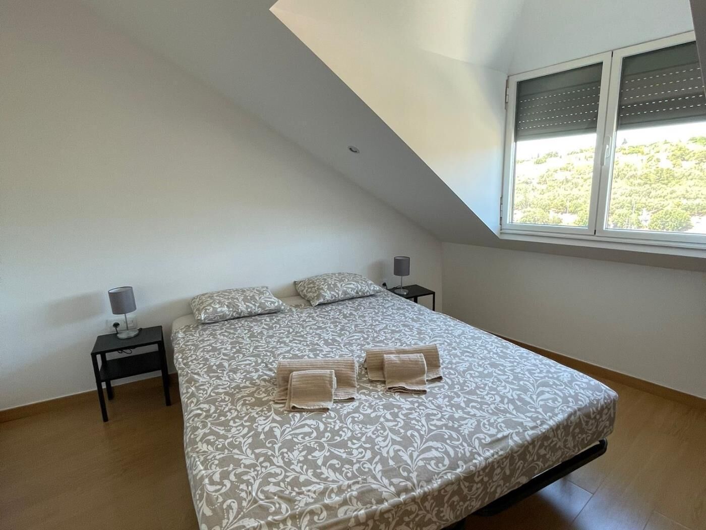 Een slaapkamer met een schuine kap, een groot bed met behang, twee nachtkastjes met lampen, en een raam met jaloezieën.