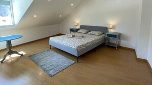 Un dormitorio con techo inclinado, una cama con ropa de cama estampada, dos mesitas de noche con lámparas, una pequeña mesa con una silla y una ventana que proporciona luz natural.