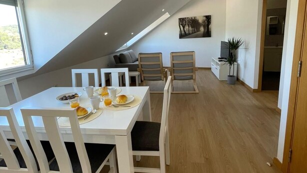 Un comedor moderno con una mesa blanca dispuesta para el desayuno, incluyendo croissants y jugo de naranja. Se observan dos sillas de madera y un sofá al fondo.