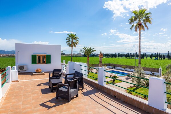 Une terrasse avec mobilier d’extérieur, une piscine et une vue sur un champ verdoyant et des palmiers.