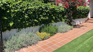 Un sendero en un jardín con piso de baldosas, bordeado por plantas verdes y una pared blanca.