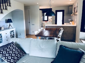 A modern living room with a sofa, a dining table, and a kitchen area with a wooden cabinet and white chairs.