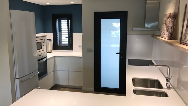 Una cocina compacta equipada con refrigerador, microondas y horno empotrado. Se aprecia una ventana con persianas, una puerta y un lavamanos con grifo.