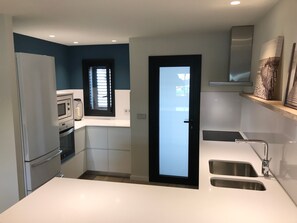 A compact kitchen with a refrigerator, microwave, and built-in oven. There's a window with blinds, a door, and a sink with a faucet.