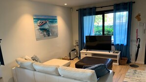 Una sala de estar moderna con un sofá blanco, un televisor de pantalla plana, un cuadro de un barco y un ventanal con cortinas azules.