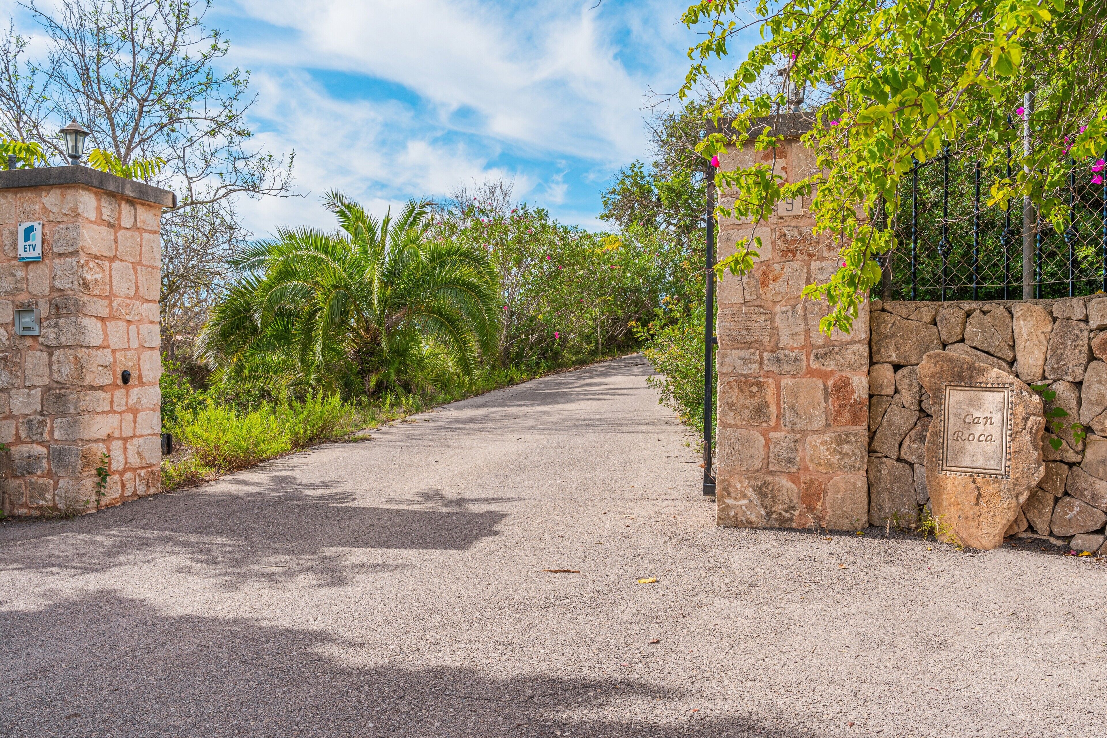 A paved pathway leading to a stone gate with a sign that reads 'Can Rocas.'