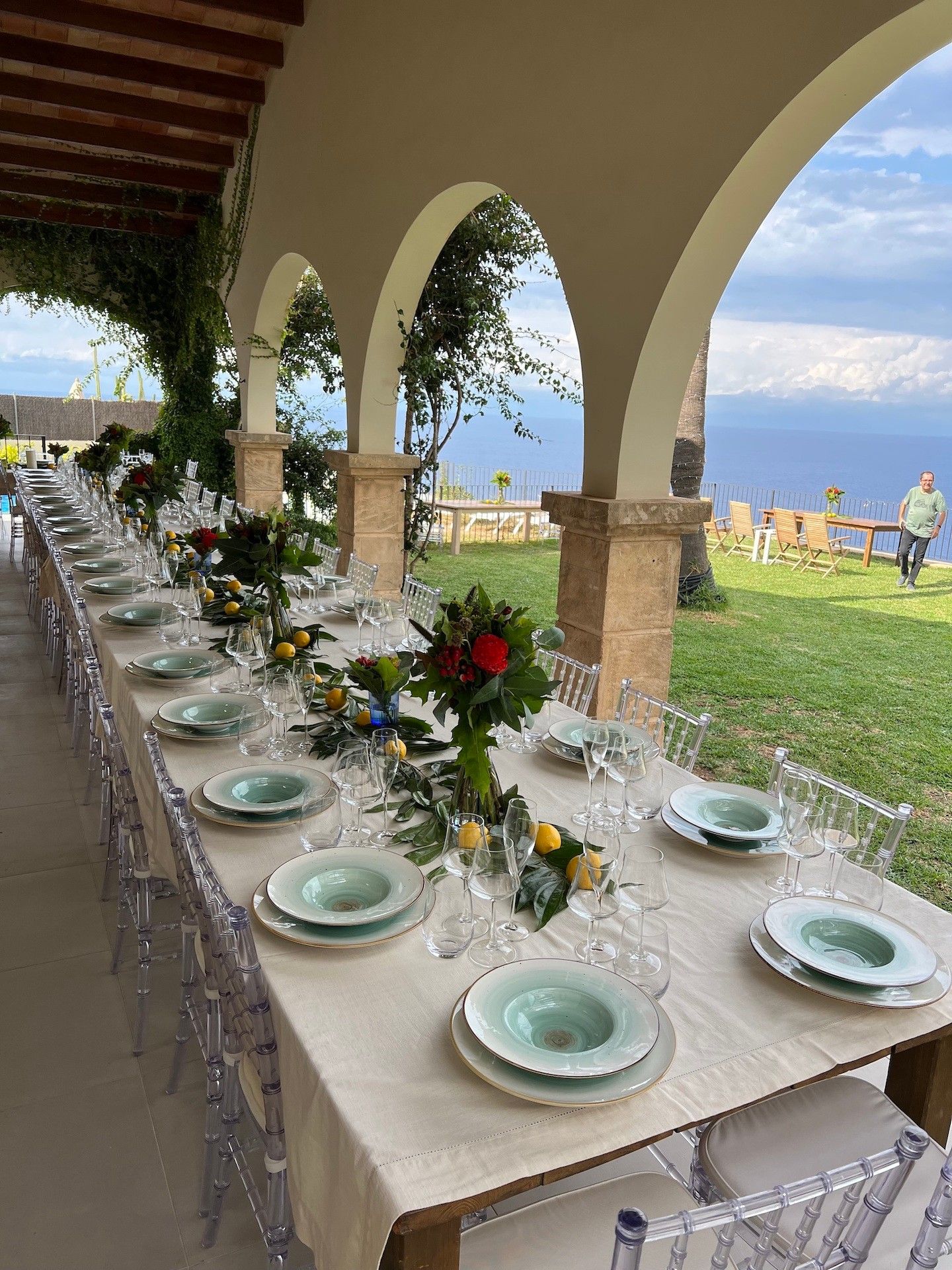 Una larga mesa de comedor al aire libre, con copas y platos de vidrio transparente, bajo una zona cubierta con pilares de piedra.
