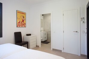 A hotel room with a bed, a chair, a small table, a bathroom with a toilet and sink, and a door leading to another room.