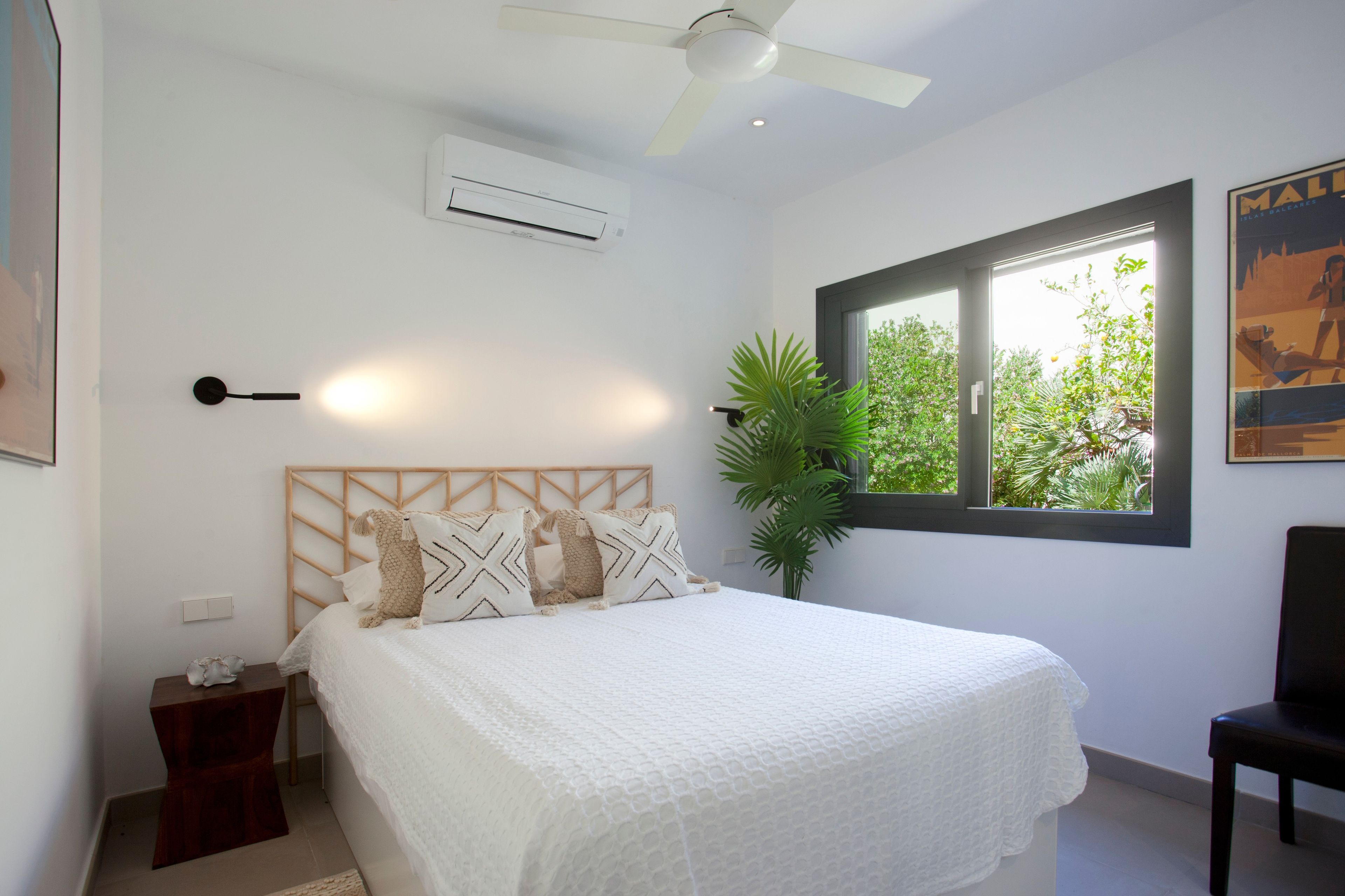 Un dormitorio con cama, ventilador de techo, aire acondicionado, ventana con vistas a la vegetación y una silla.