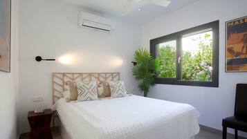 Un dormitorio con cama, ventilador de techo, aire acondicionado, ventana con vistas a la vegetación y una silla.